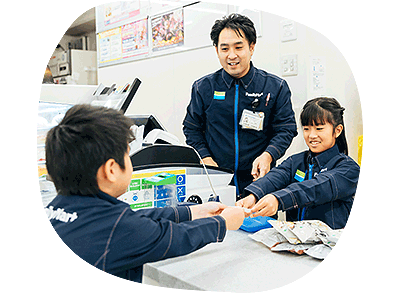 ファミリーマートレジカウンターにて職業体験をする女の子とそれを見守るファミリーマートスタッフ。